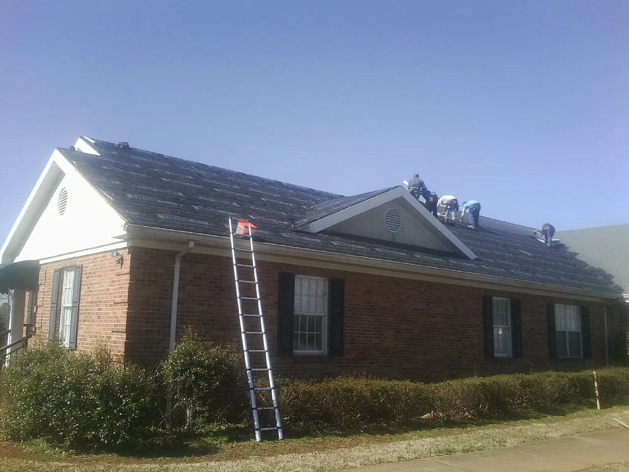 Crew tearing shingles off a brick ranch roof with a ladder set — Emergency Roof Repair work by our crew in Normangee