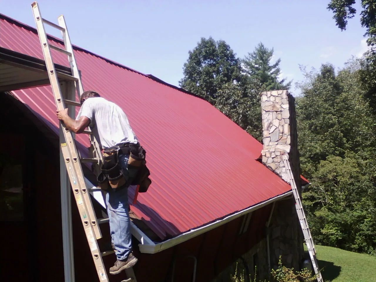 Roofer on a ladder beside a red metal roof and stone chimney — Soffit & Fascia Repair by Roofing Brothers in Normangee